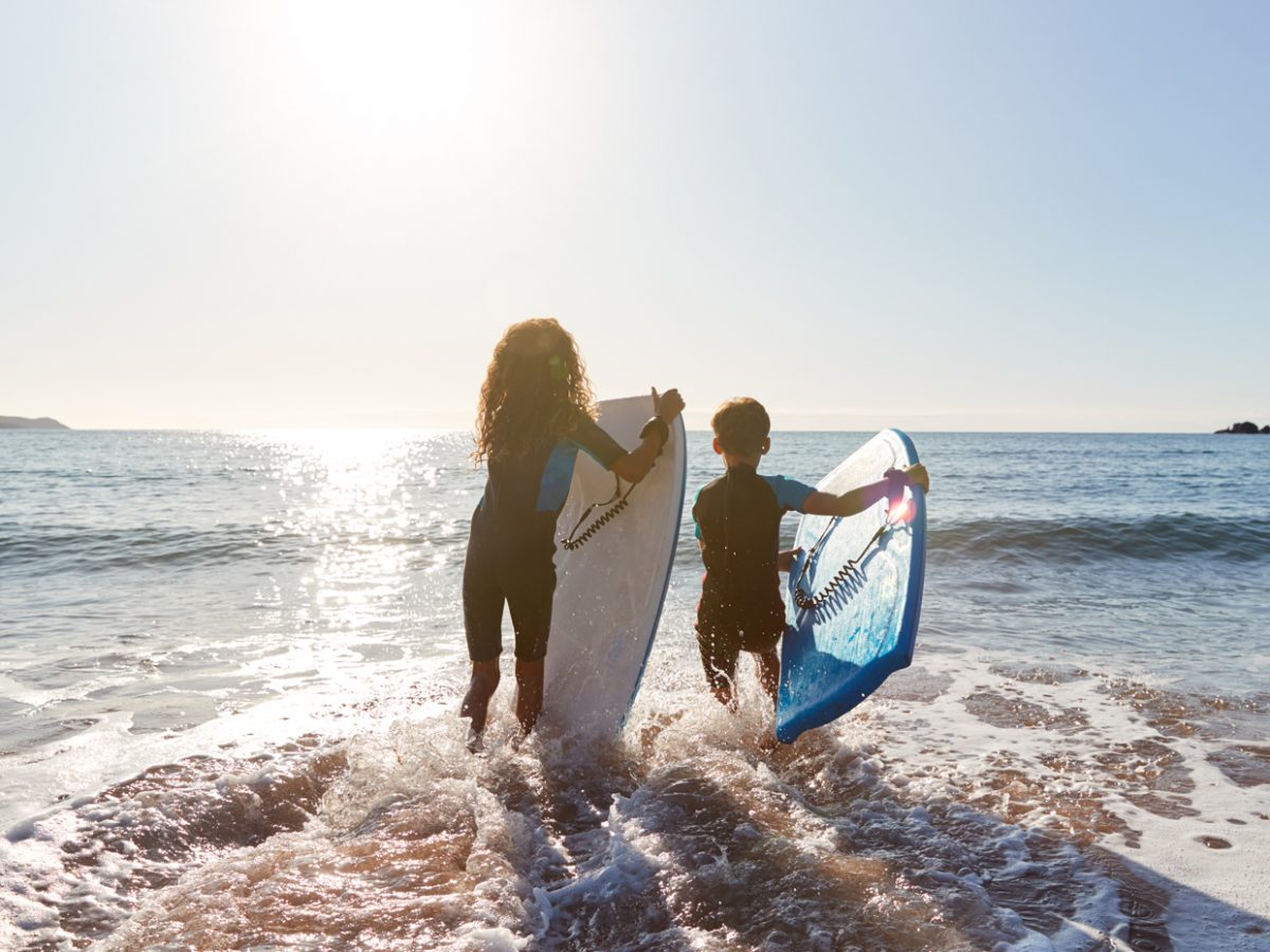 Rear View Of Two Children Wearing Wetsuits Running Into Sea With Bodyboards On Cornwall Vacation